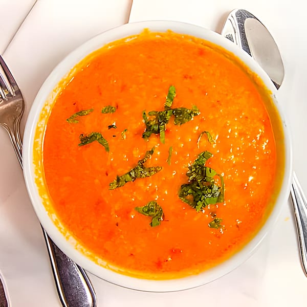 Overhead view of a white bowl filled with vibrant orange Indian tomato soup, garnished with fresh herbs.