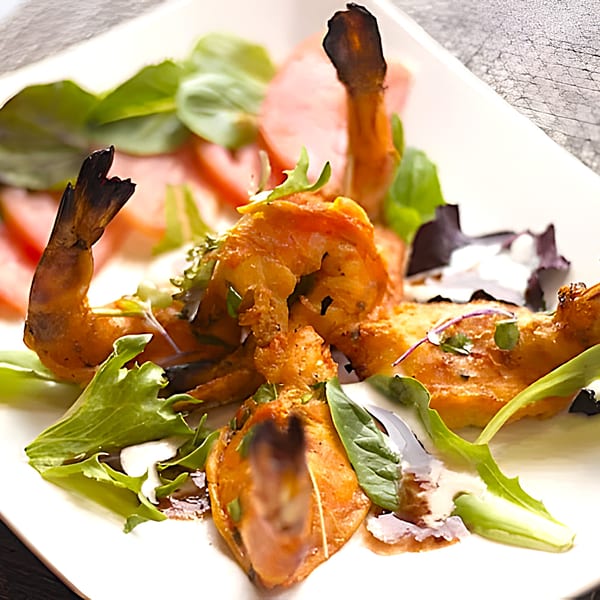 Indian-spiced shrimp with charred tails, served on a white plate with fresh greens and tomato slices.